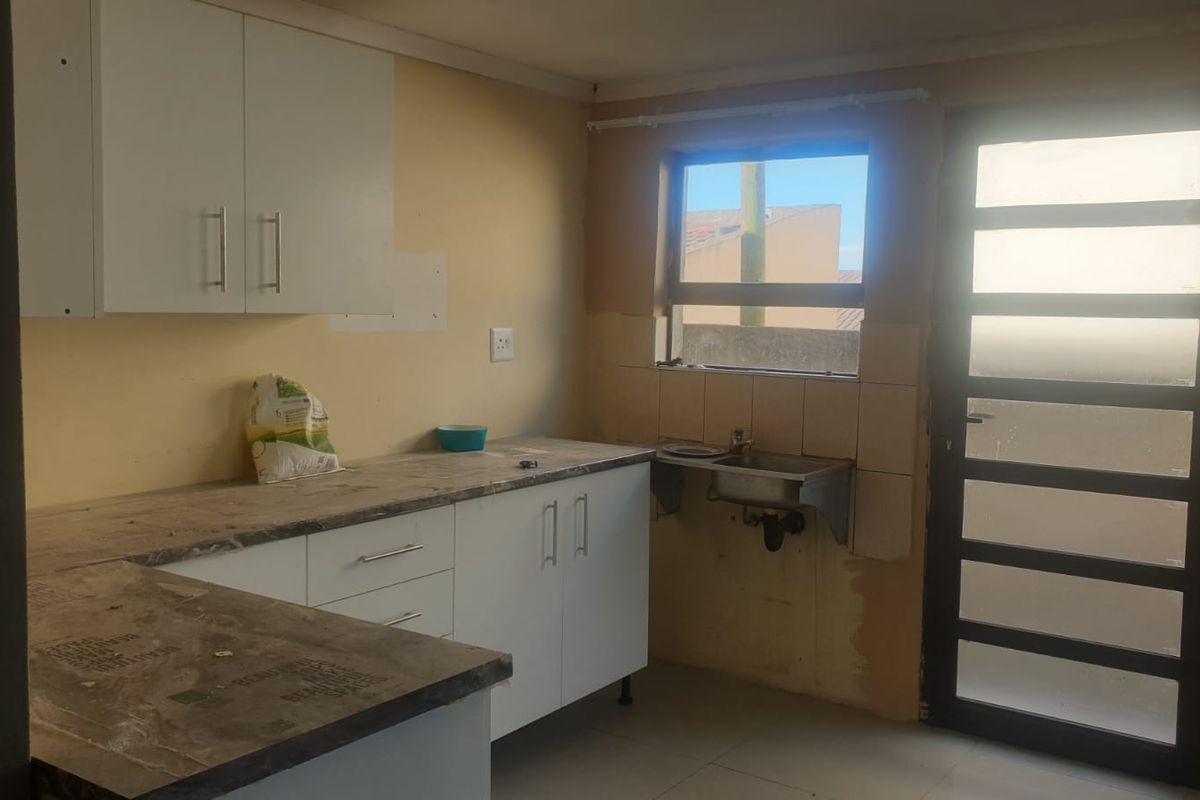 A functional kitchen area featuring white cabinetry, laminate countertops, and a stainless steel sink, currently requiring finishing touches to the walls and tiling.