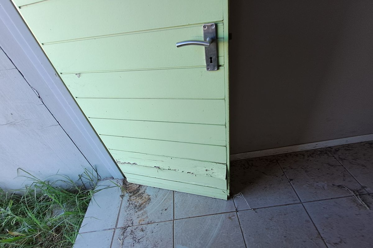 Close-up view of a weathered exterior door showing significant wood rot at the base and peeling paint, situated on a tiled patio area.