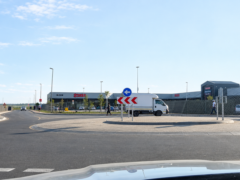 A view of a paved roundabout and access road leading to a commercial shopping center with modern retail buildings.