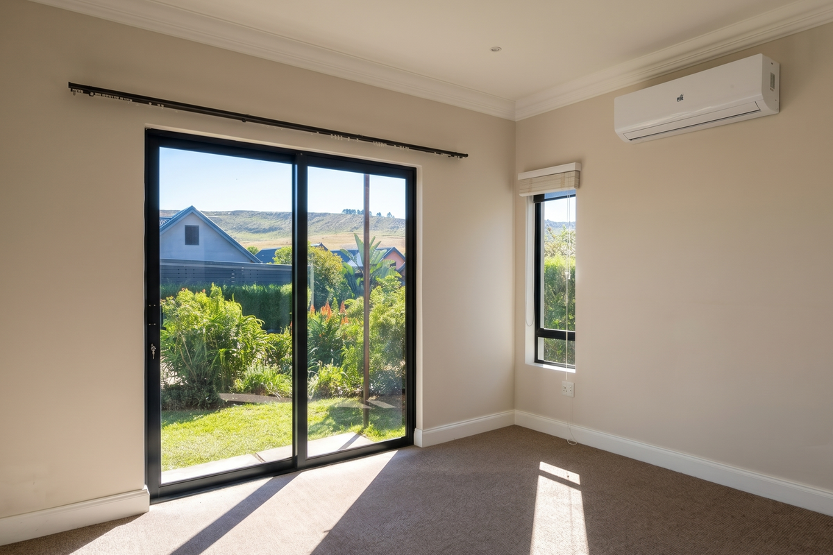 A bright, empty bedroom featuring neutral walls, carpet flooring, a split-system air conditioner, and large sliding glass doors opening to a garden view.