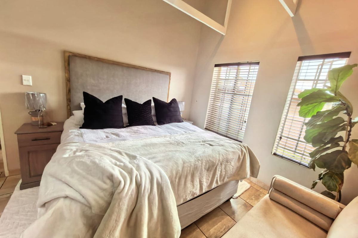 A bright bedroom featuring a vaulted ceiling with exposed rafters, tiled flooring, and dual windows fitted with venetian blinds.
