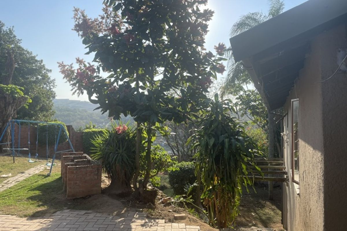 A rear garden area featuring paved sections, established trees, and a view of the surrounding landscape, adjacent to the house exterior.