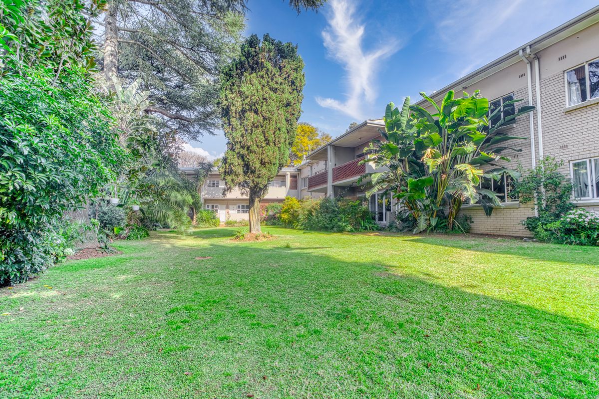 A view of the rear exterior of the apartment complex, featuring a well-maintained communal garden, mature trees, and private balconies.