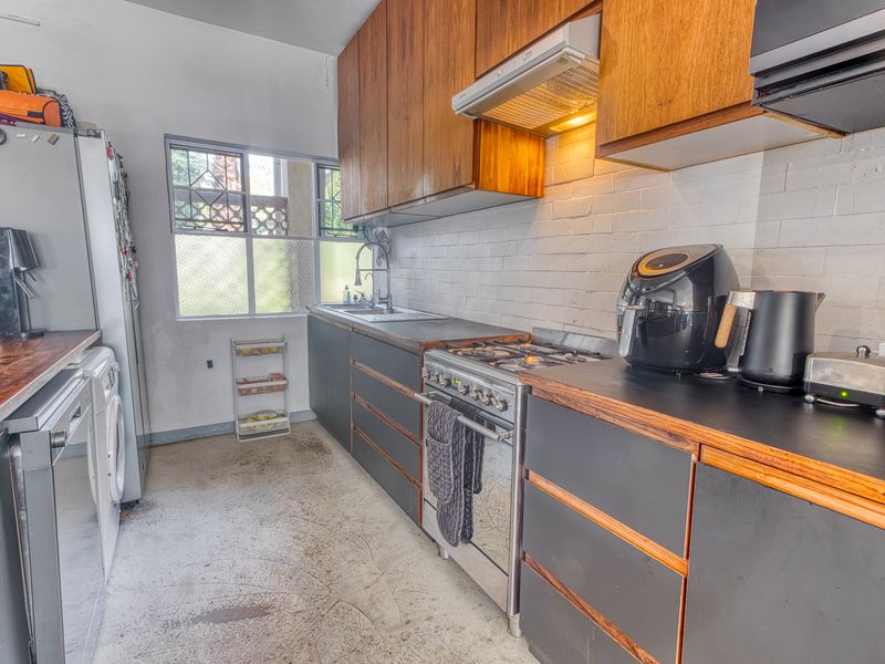 A compact kitchen featuring dark cabinetry, timber countertops, a gas stove with extractor fan, and exposed brick walls, requiring some cosmetic updates.