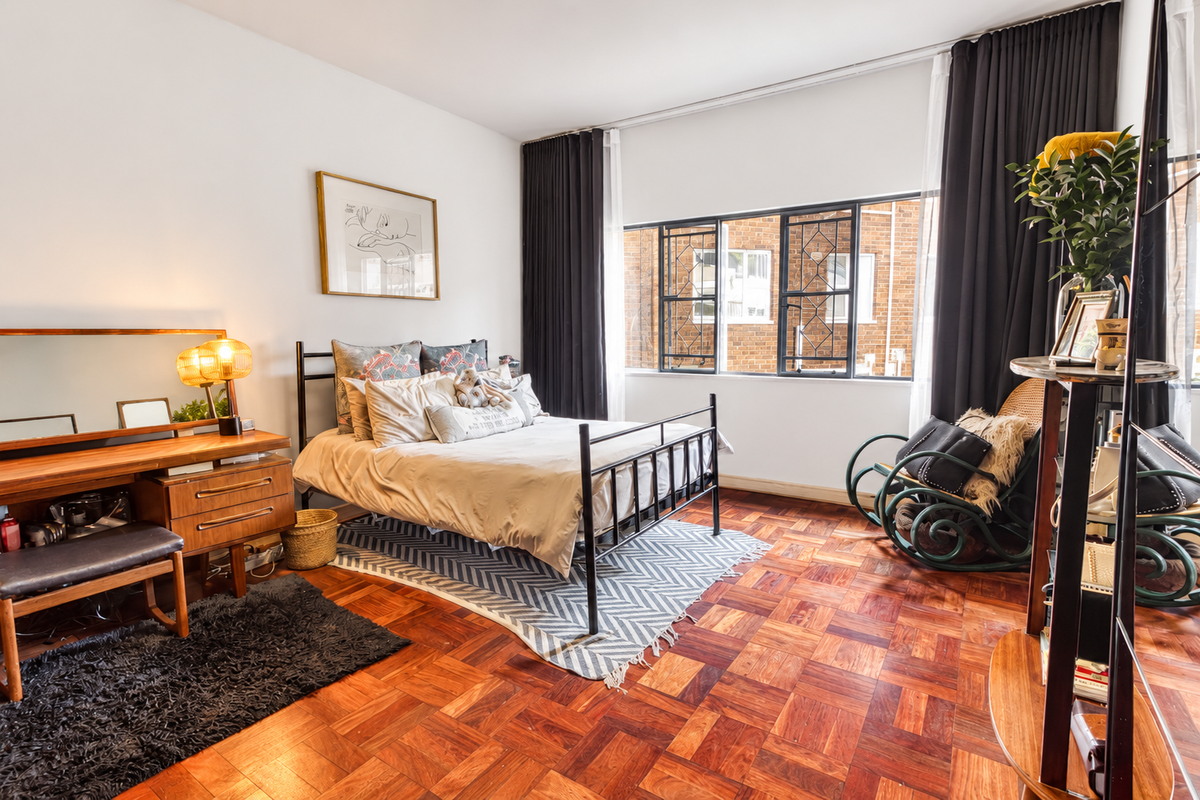 A well-lit bedroom featuring classic parquet flooring and large metal-framed windows with integrated curtain tracks.