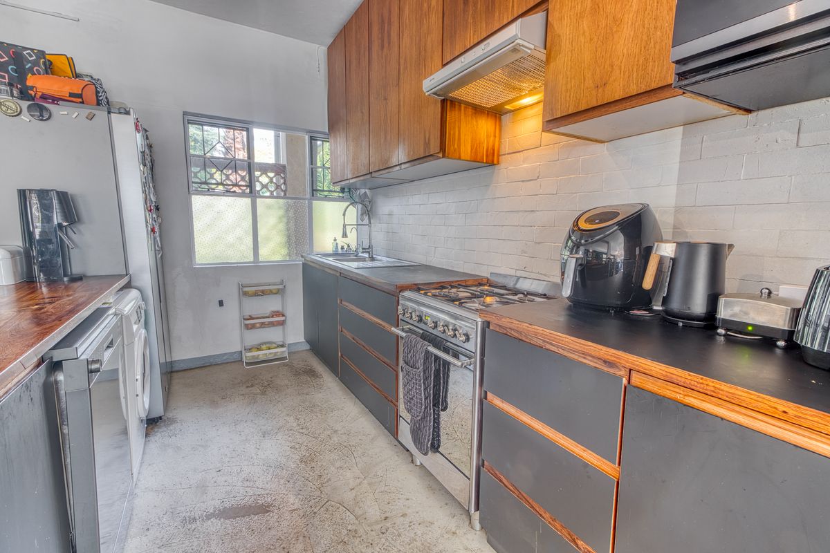 A compact kitchen featuring dark cabinetry, timber countertops, a gas stove with extractor fan, and exposed brick walls, requiring some cosmetic updates.