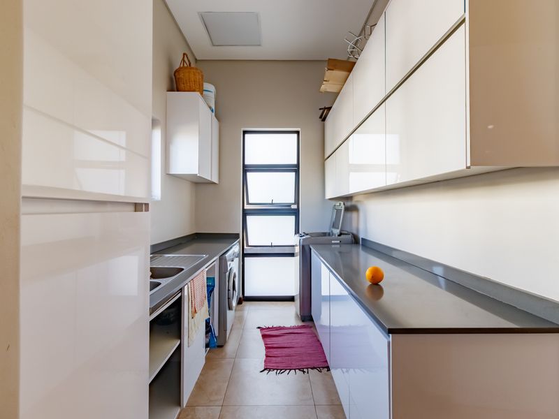 A modern, functional laundry room featuring sleek white cabinetry, stone countertops, a stainless steel sink, and tiled flooring with natural light.