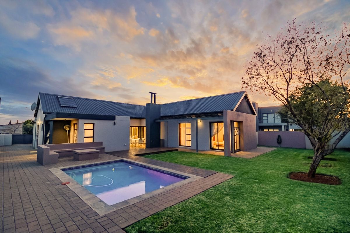 Rear view of a modern home featuring a paved patio, swimming pool, and well-maintained garden under a sunset sky.