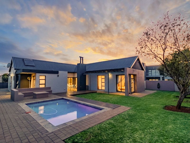 Rear view of a modern home featuring a paved patio, swimming pool, and well-maintained garden under a sunset sky.