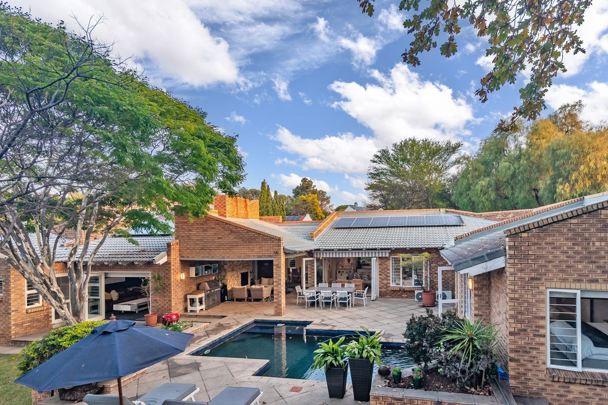 Spacious rear exterior featuring a paved patio, covered entertainment area with built-in braai, and a swimming pool set against a brick-faced home.