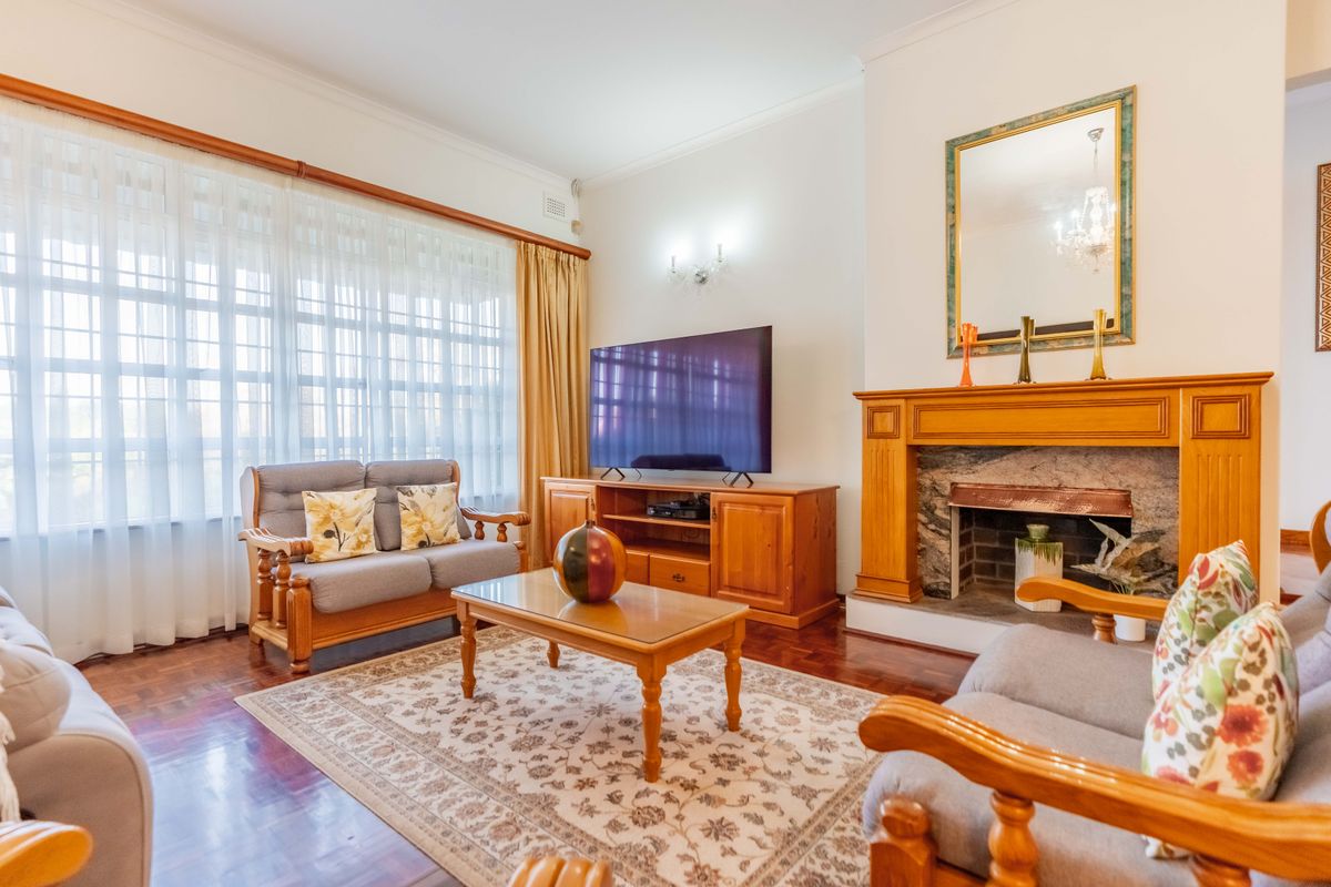 A comfortable living room featuring classic parquet flooring, a prominent wooden fireplace mantel with stone surround, and large windows with natural light.
