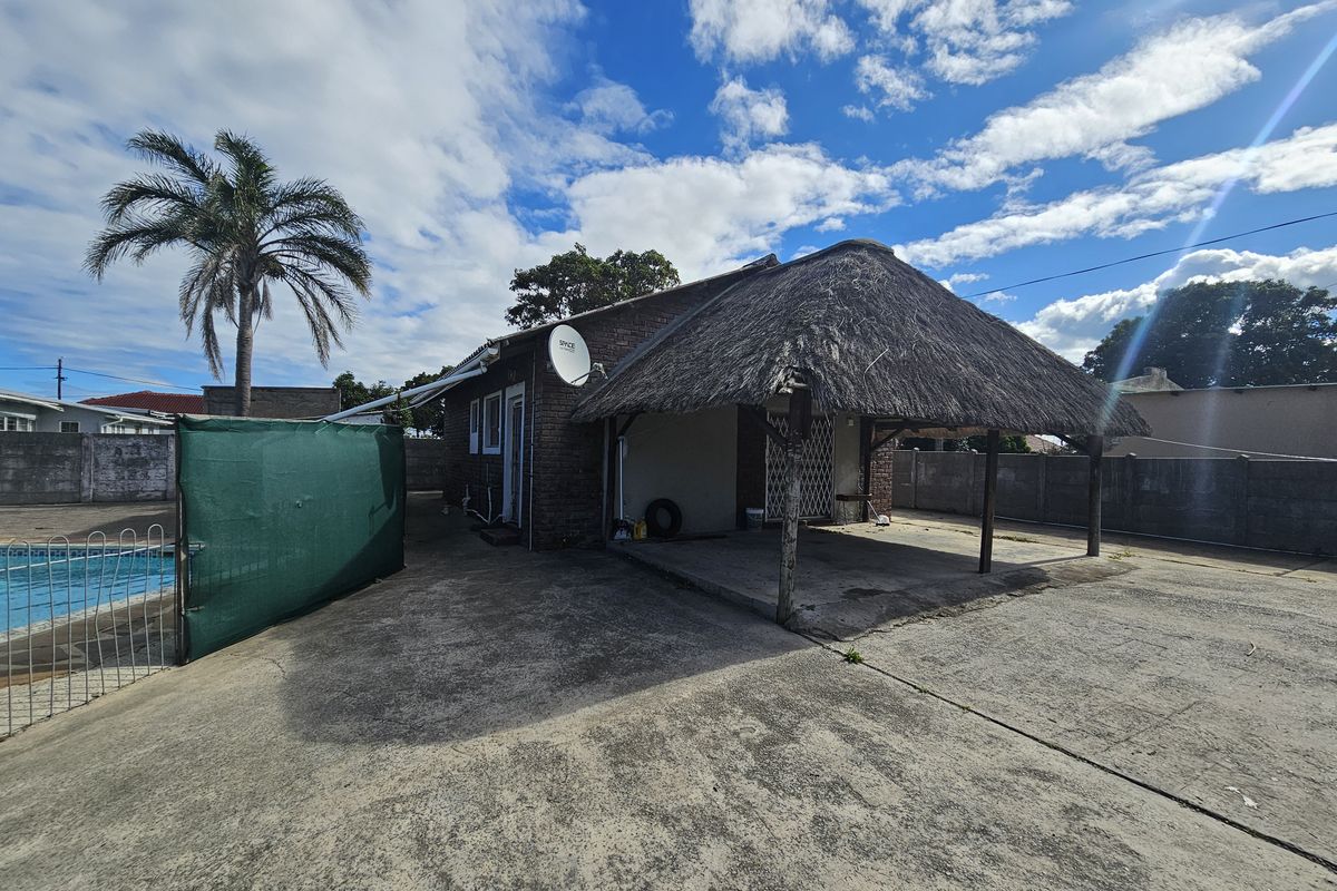Rear exterior view featuring a thatched roof patio area, concrete paving, and an adjacent swimming pool with perimeter walling.