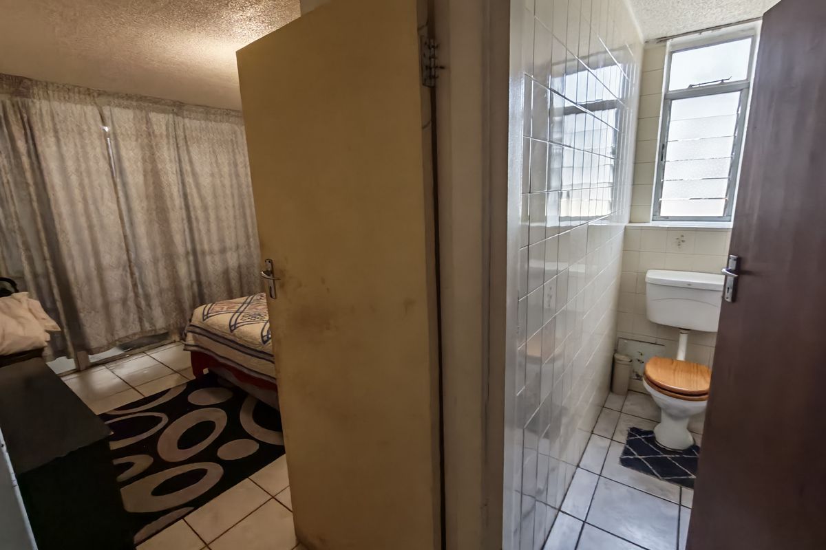A dated bathroom featuring ceramic wall and floor tiles, a standard toilet, and a louvered window, showing signs of wear on the door.