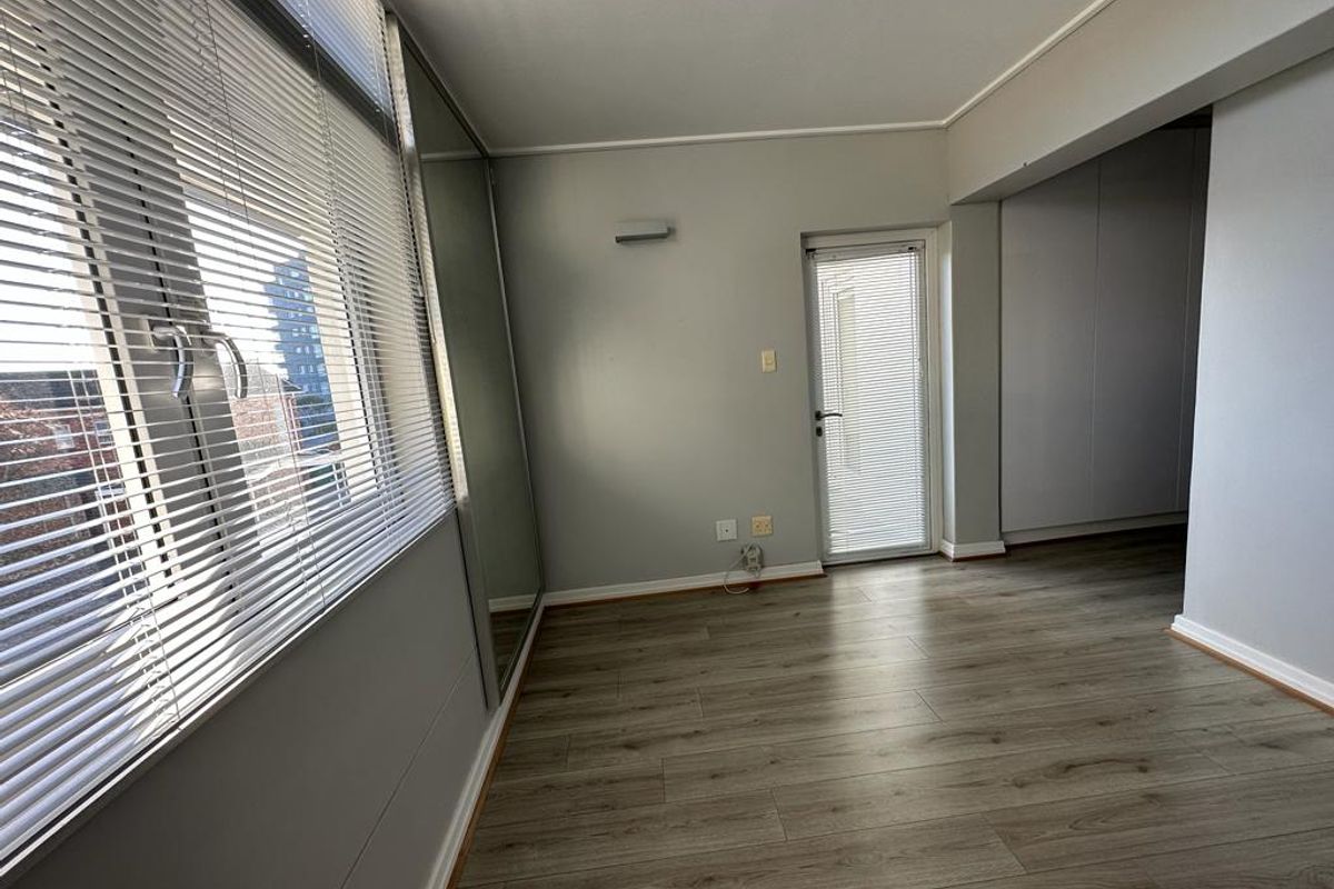 A bright, empty bedroom featuring modern laminate flooring, built-in cupboards, and large windows with venetian blinds, offering a clean and neutral living space.