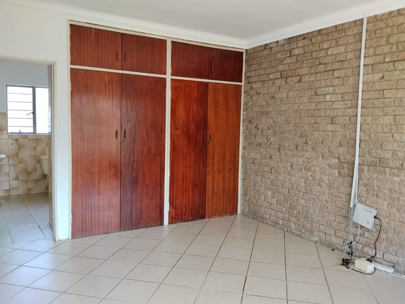 A bedroom featuring built-in wooden cupboards, an exposed brick feature wall, and tiled flooring, with an adjacent bathroom visible through the doorway.