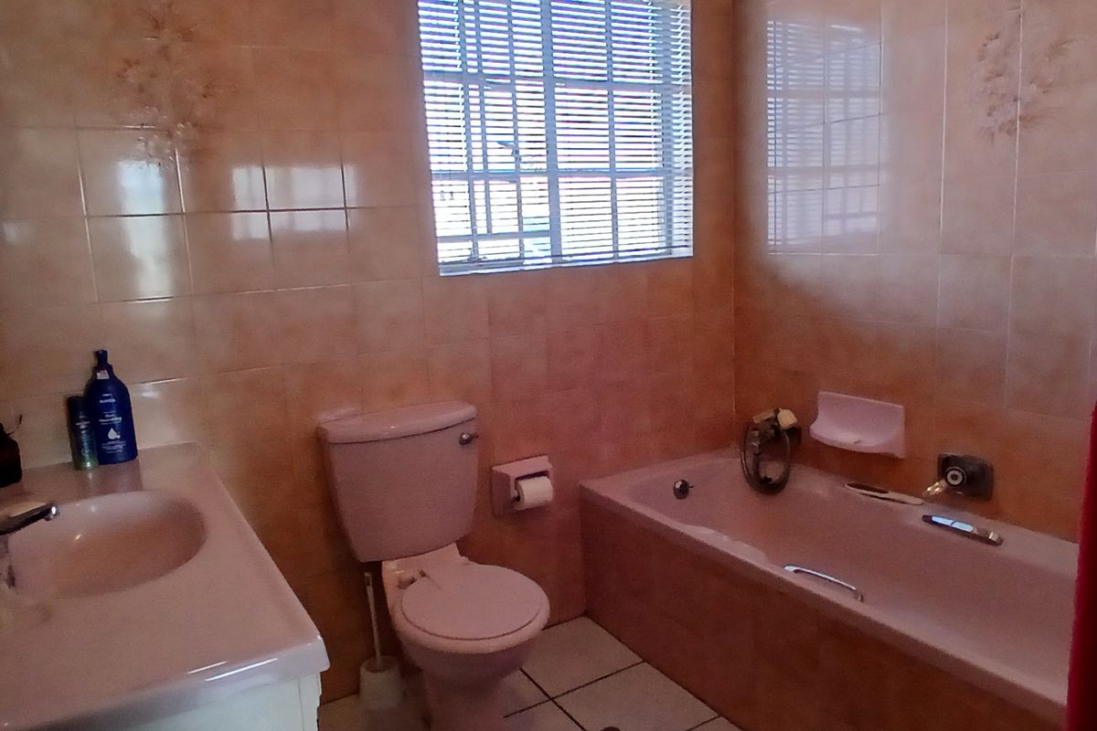 A dated bathroom featuring a pink bathtub, matching toilet, and vanity basin with full-height wall tiling and a window with fitted blinds.