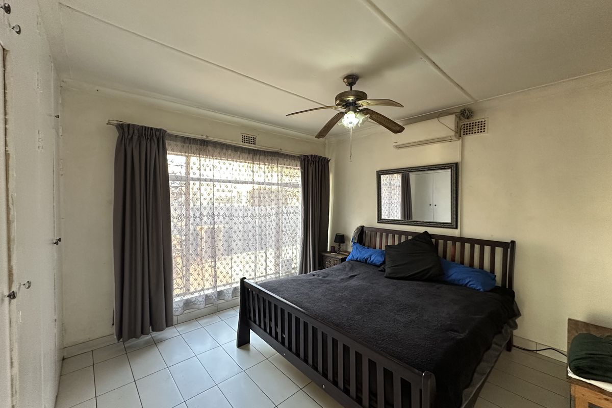 A bedroom featuring tiled flooring, a ceiling fan, and a wall-mounted air conditioning unit, with large windows providing natural light.