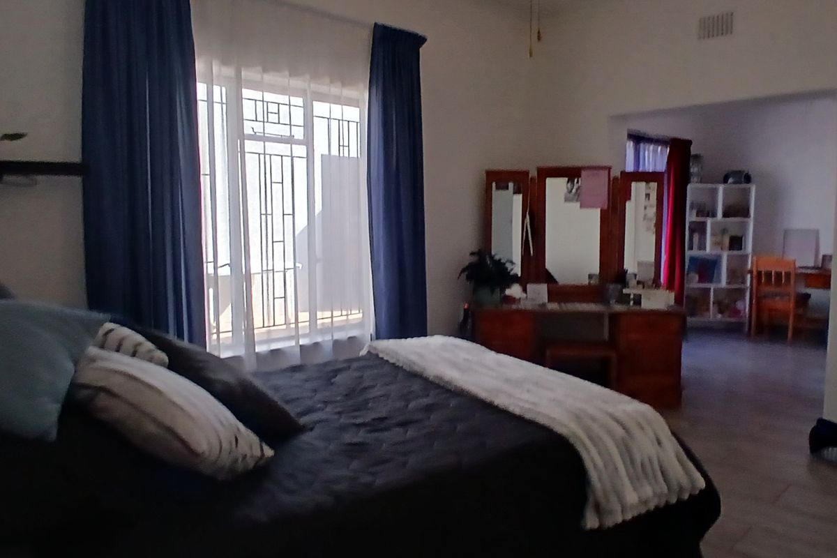 A spacious bedroom featuring tiled flooring, large windows with security bars, and an open-plan layout leading into an adjacent room.