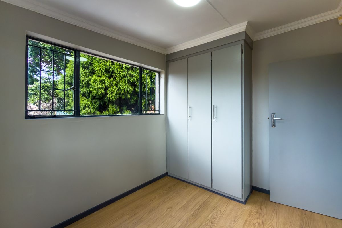 A bright bedroom featuring light-toned laminate flooring, built-in cupboards, and a window with security bars overlooking the garden.