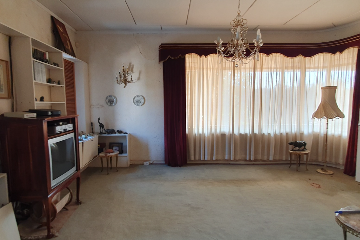 A spacious living room featuring built-in shelving, a large window with decorative curtains, and a central chandelier, requiring cosmetic updates to modernize the space.