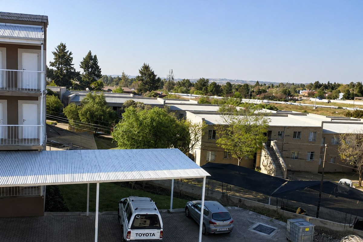 An elevated view of the apartment complex grounds, featuring paved parking areas, covered carports, and surrounding residential buildings with metal roofing.