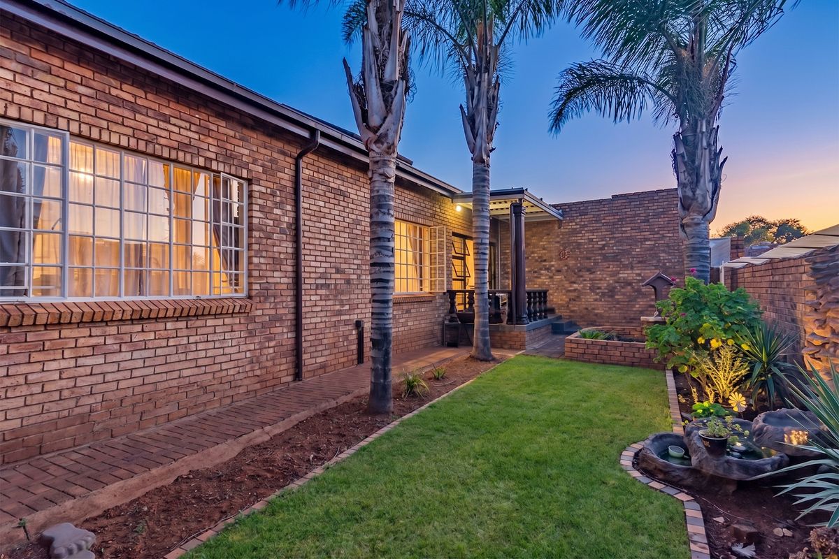 A well-maintained rear exterior view featuring a face brick facade, a paved walkway, a covered patio, and a landscaped garden with a water feature.