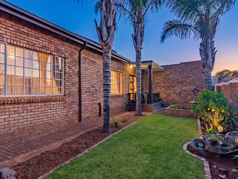 A well-maintained rear exterior view featuring a face brick facade, a paved walkway, a covered patio, and a landscaped garden with a water feature.