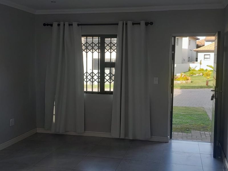 An empty living room featuring neutral tiled flooring, security bars on the window, and direct access to the exterior garden area.