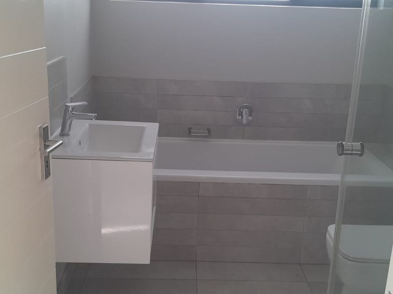 A modern, clean bathroom featuring a wall-mounted vanity, built-in bathtub, and a glass shower screen with neutral grey wall and floor tiling.