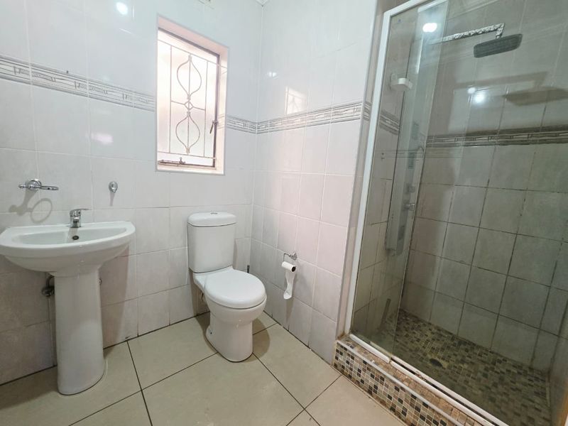 A functional bathroom featuring a pedestal washbasin, toilet, and a glass-enclosed shower with tiled finishes and a security window grille.