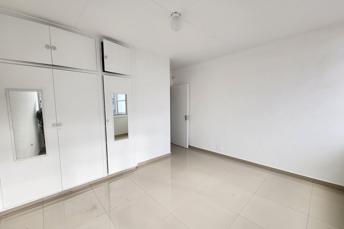 A bright, empty bedroom featuring white built-in cupboards with mirrored accents and neutral tiled flooring throughout.
