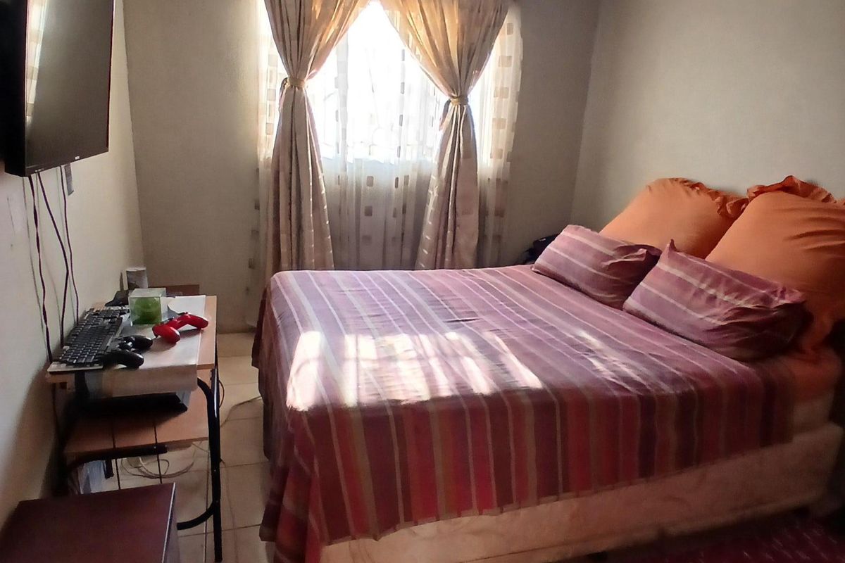A compact bedroom featuring tiled flooring and a window with curtain rails, showing signs of wear on the ceiling.