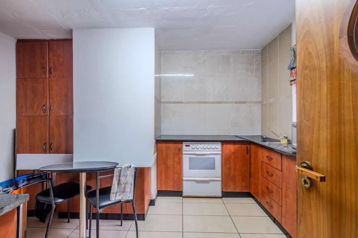 A compact kitchen featuring wood-finish cabinetry, a built-in oven with hob, dark countertops, and neutral floor tiling.