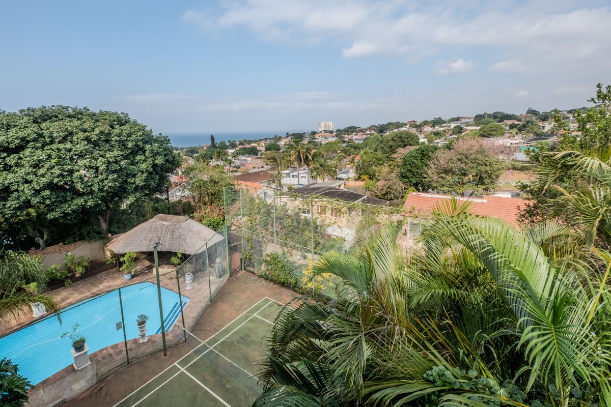 Elevated view of a private residential property featuring a swimming pool, thatched gazebo, and a fenced tennis court set within a lush garden.