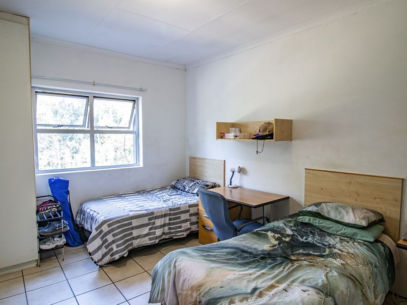 A functional bedroom featuring tiled flooring, built-in cupboards, and an aluminium window frame, providing a practical living space with neutral finishes.