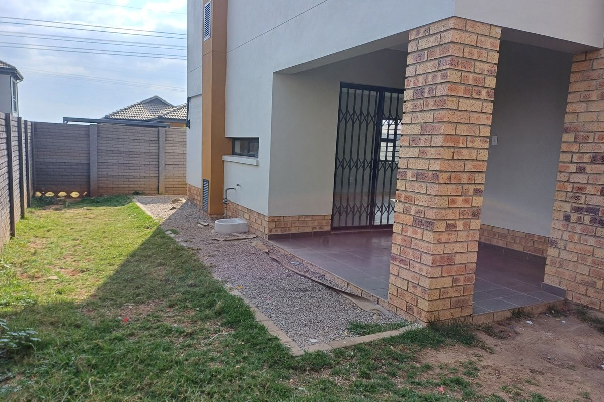 A rear view of the apartment featuring a covered patio with brick pillars, tiled flooring, security gate, and an adjacent garden area.