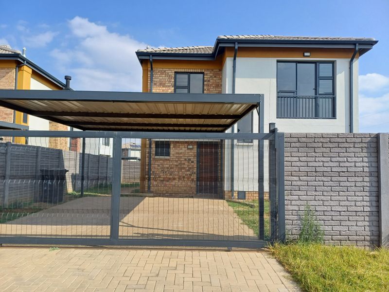 Front elevation of a modern two-story apartment featuring a paved driveway, covered carport, brick and plaster facade, and secure perimeter fencing.