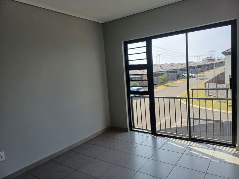 A bright, empty room featuring neutral tiled flooring and a large sliding glass door that opens onto a balcony with a neighbourhood view.