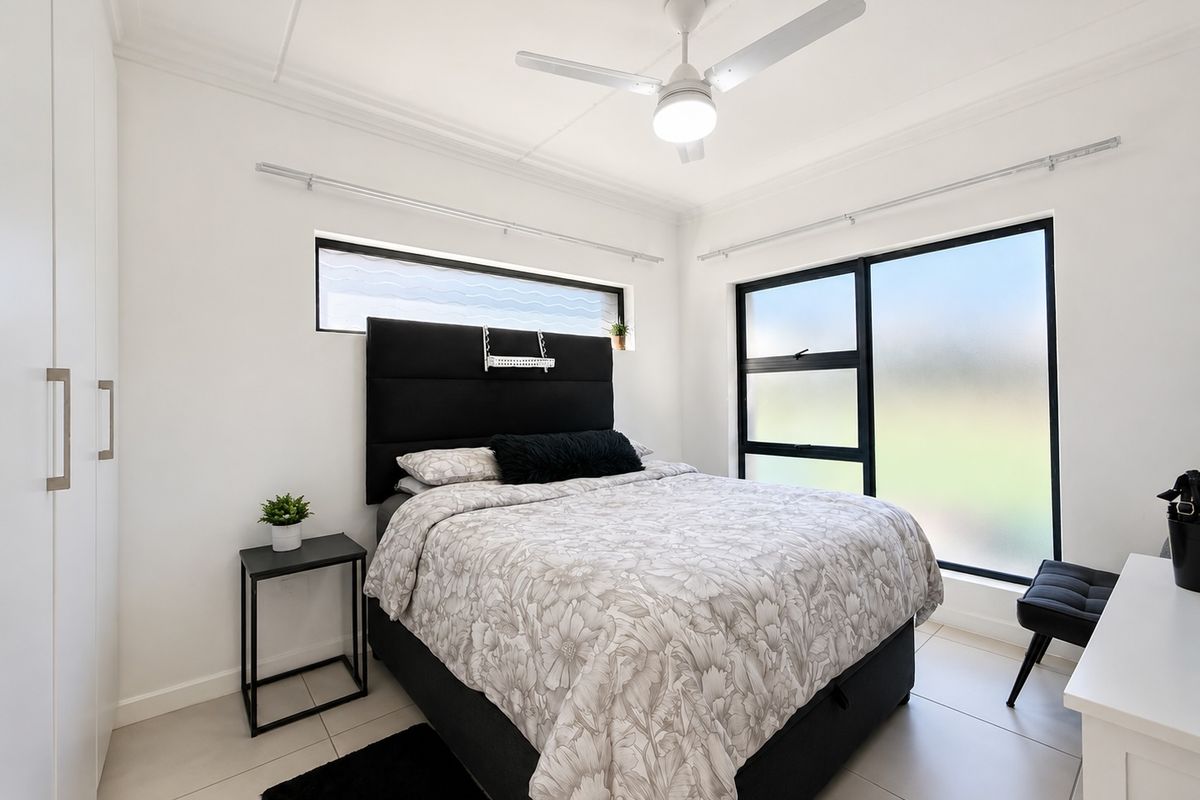 A modern, well-lit bedroom featuring tiled flooring, a ceiling fan, and built-in wardrobe storage with large windows providing natural light.
