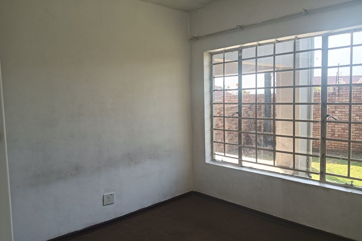 An empty bedroom featuring carpet flooring and a large window with security bars, showing signs of wear on the walls and flooring.