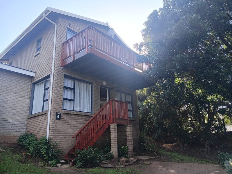 Rear view of a two-story brick cottage featuring a timber balcony, external staircase, and established garden surroundings.