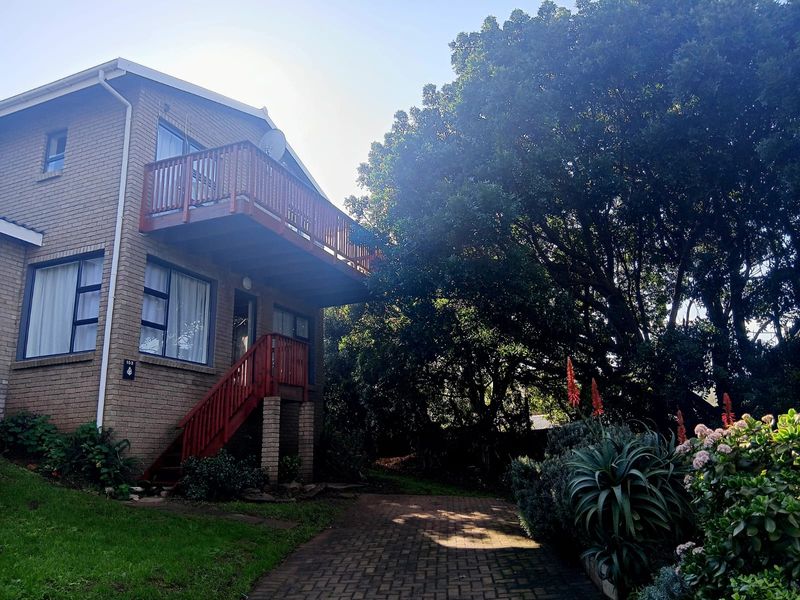 Rear view of a brick cottage featuring an elevated wooden balcony, external staircase, and a paved driveway surrounded by mature trees and garden landscaping.