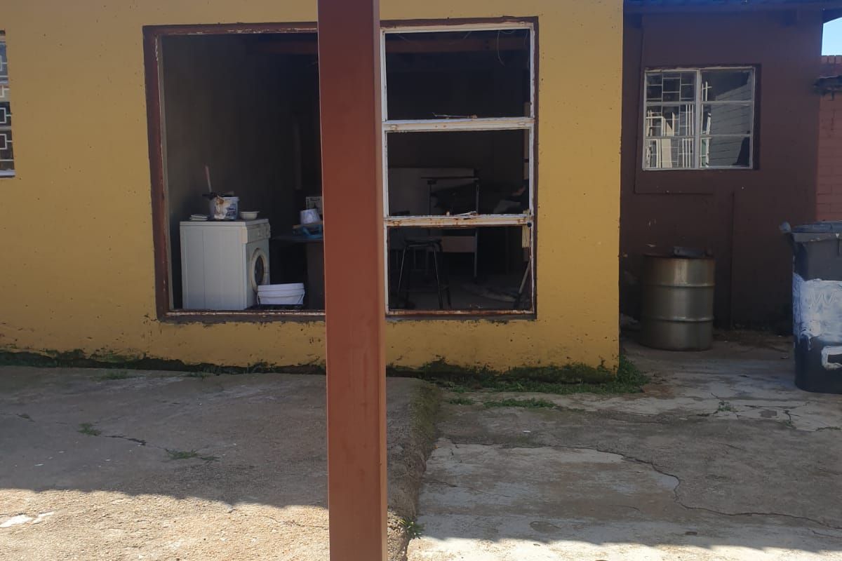 Rear exterior view of a property showing a concrete patio area and a building structure with missing window panes and weathered exterior finishes.