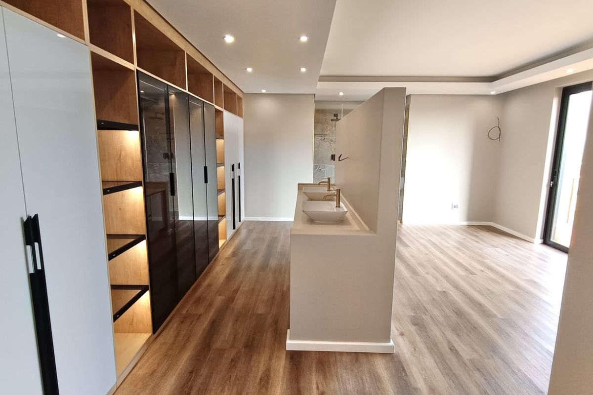 Modern open-plan bedroom suite featuring extensive built-in cabinetry, a central double vanity partition, and high-quality laminate flooring with recessed lighting.
