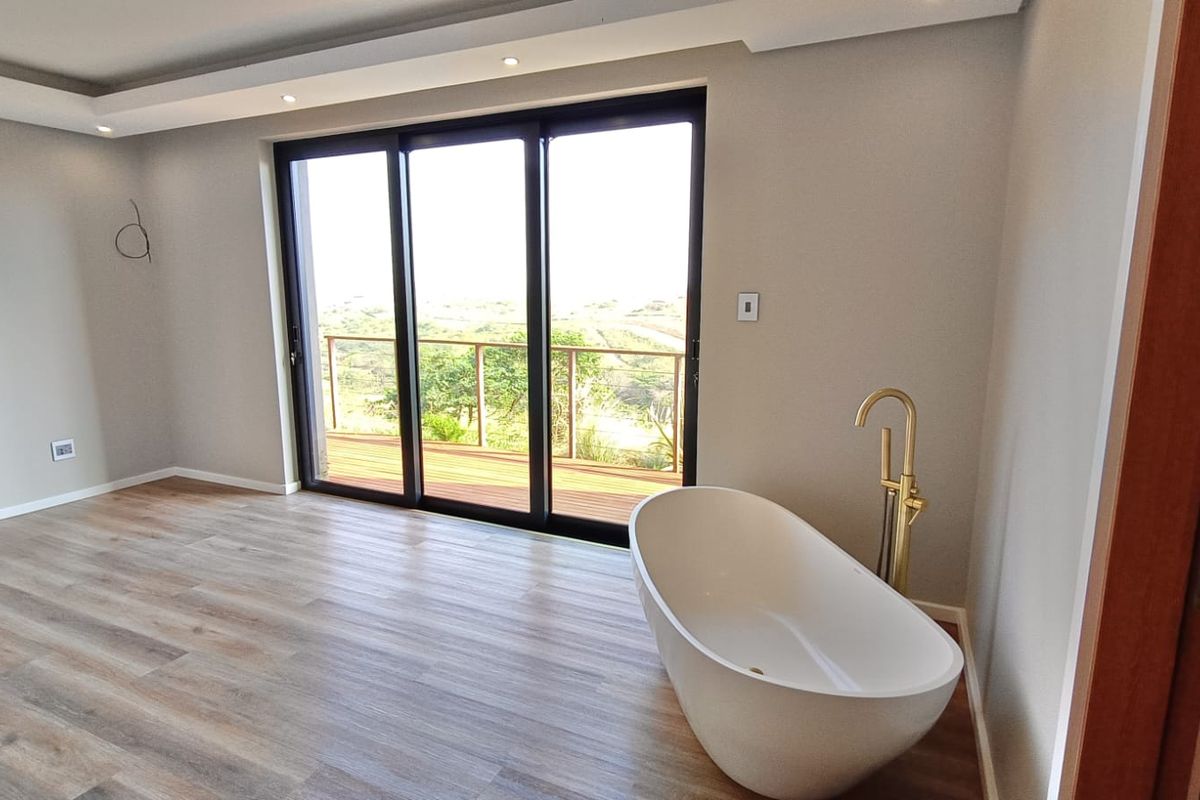 Modern open-plan bathroom featuring a sleek freestanding bathtub with gold fixtures, wood-look flooring, and large sliding doors opening to a scenic outdoor deck.