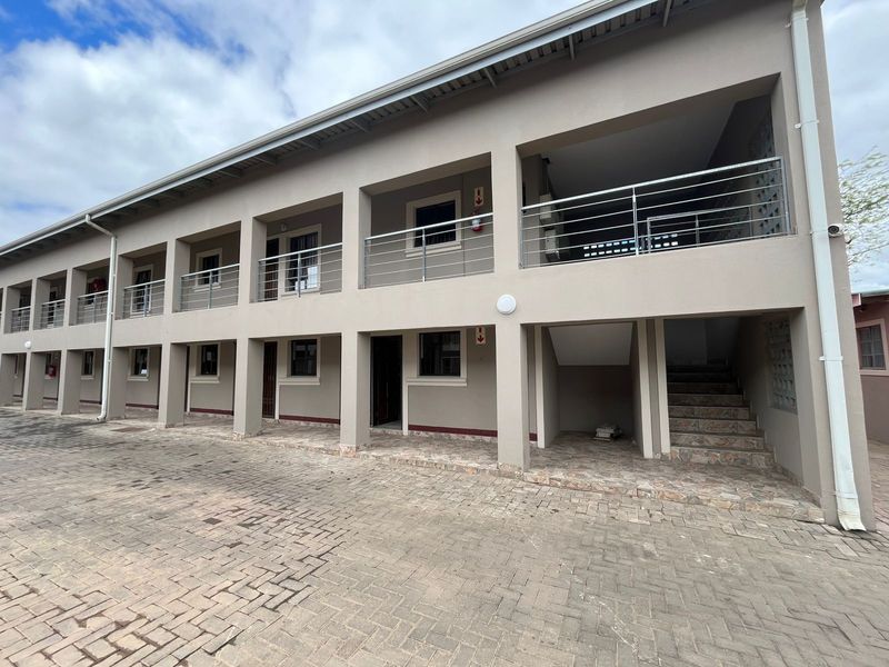 A multi-story apartment building exterior featuring a paved driveway, covered balconies with metal railings, and an external staircase.