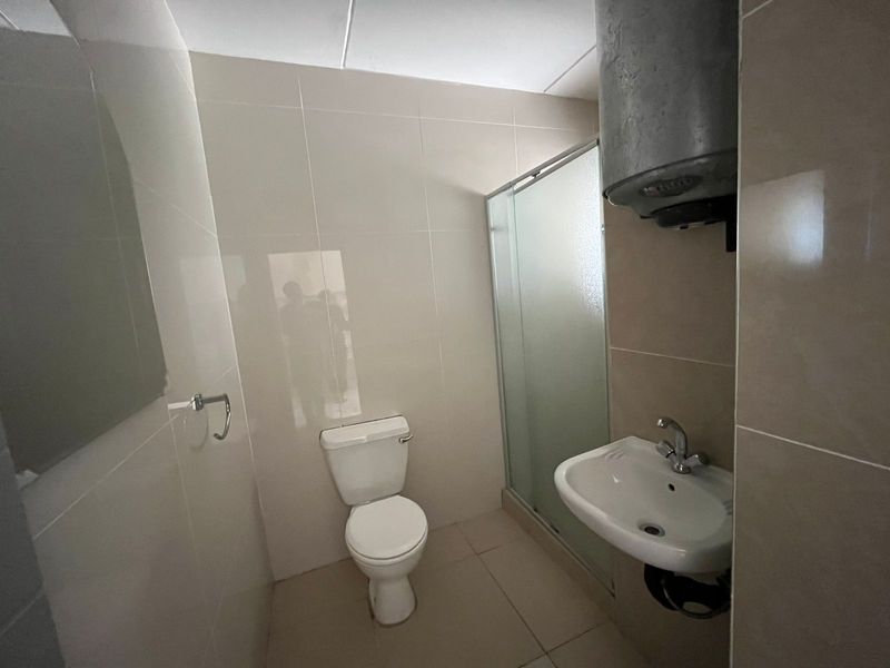 A functional bathroom featuring a shower enclosure, toilet, wall-mounted basin, and an exposed electric water heater with tiled finishes throughout.