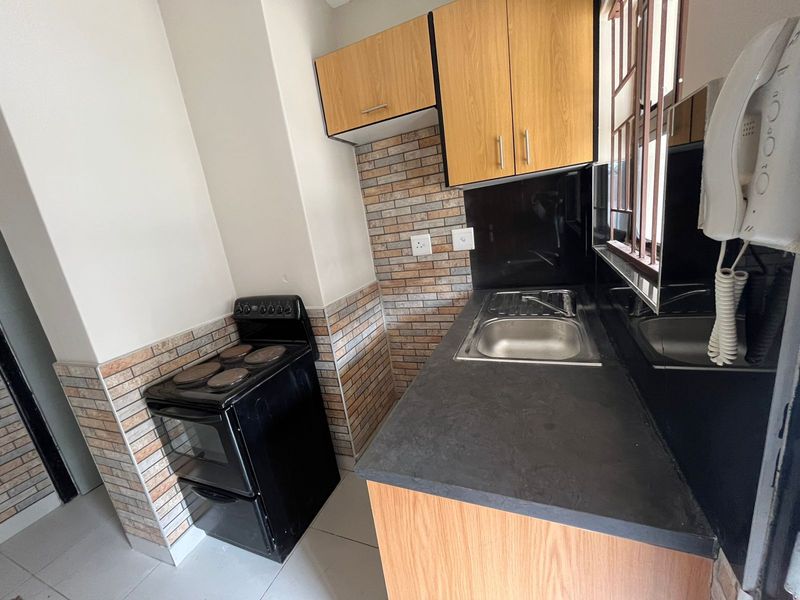 Compact kitchen featuring wood-finish cabinetry, black countertops, a stainless steel sink, and a built-in electric stove with a decorative tile backsplash.