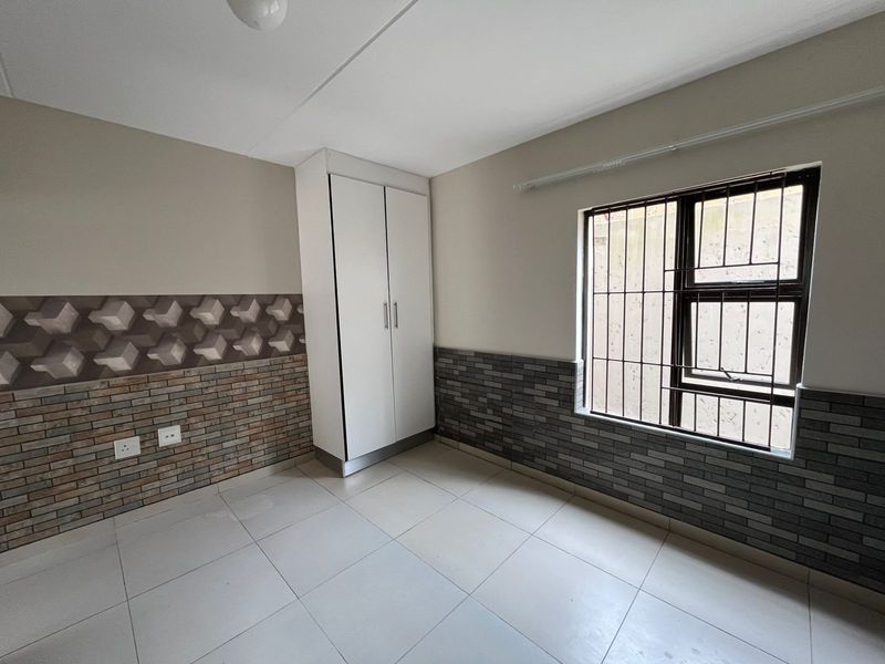A clean, empty bedroom featuring tiled flooring, a built-in wardrobe, and decorative wall cladding with security bars on the window.