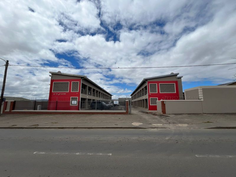 Front exterior view of a multi-story apartment complex featuring red accent walls, paved driveway access, and secure perimeter fencing with razor wire.